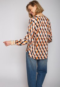 Bluzka z długim rękawem z lekkiego materiału, zdobiona abstrakcyjnym wzorem wielokolorowych owali (pomarańczowy, granatowy, jasnoniebieski) na kremowym tle.