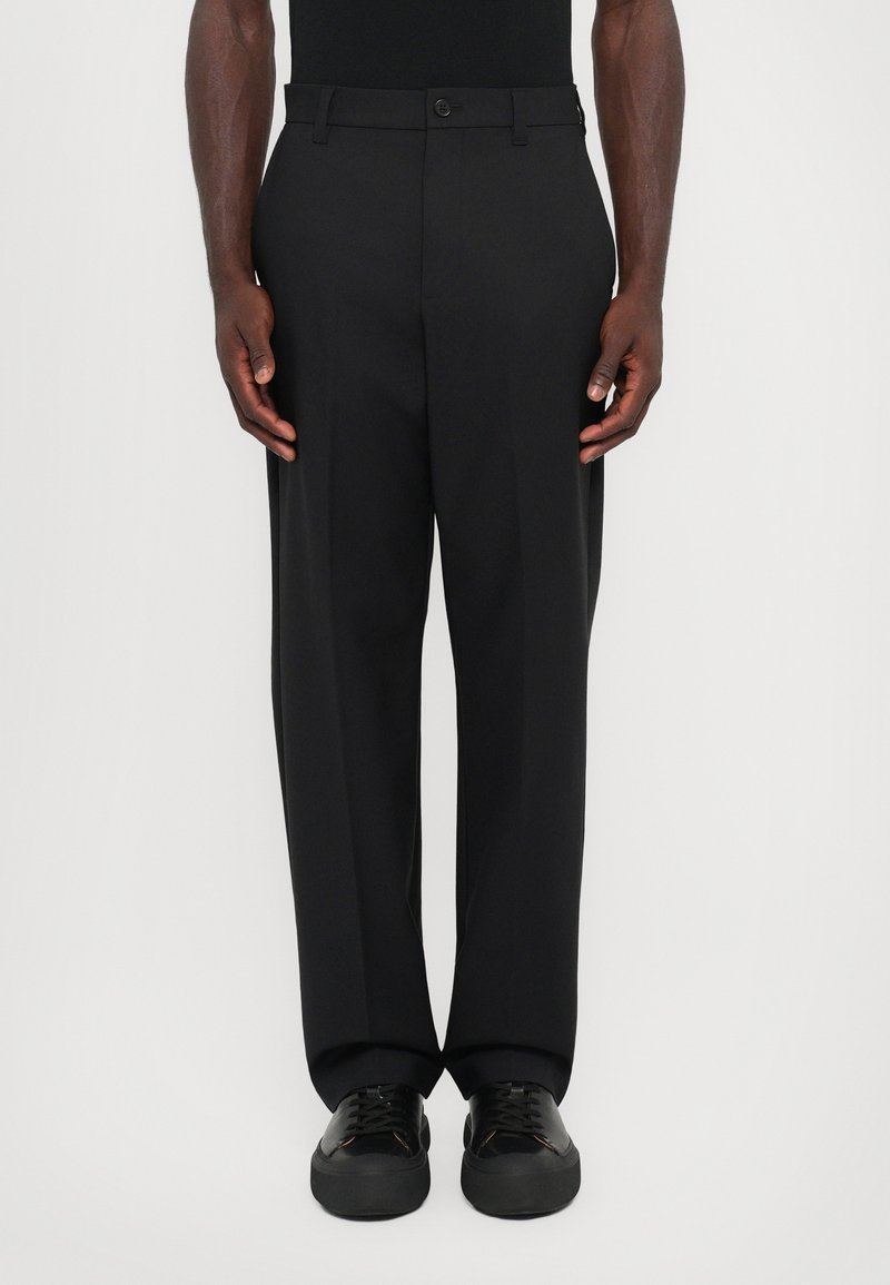 Homme portant un pantalon noir droit et des chaussures noires à lacets, debout sur un fond blanc.