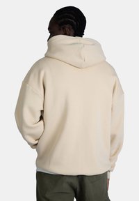 Felpa beige con cappuccio e vestibilità morbida, con spalle scese e una texture soffice. Vista posteriore, senza loghi o motivi visibili.