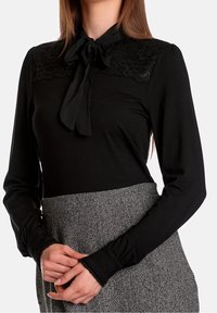 Blusa nera a maniche lunghe con colletto in pizzo, fiocco al collo e polsini aderenti. Realizzata in tessuto morbido, abbinata a una gonna grigia a vita alta con fantasia.