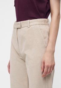 Femme portant un pantalon en velours côtelé beige avec une ceinture assortie et un haut sans manches violet foncé, main reposant le long du corps.