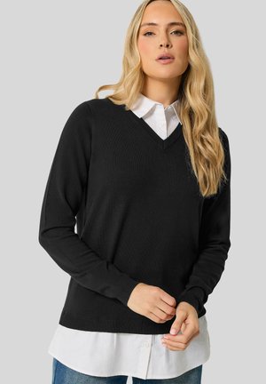 Blonde Frau trägt einen schwarzen V-Ausschnitt-Pullover über einem weißen Hemd und Jeans, steht vor einem einfarbigen hellgrauen Hintergrund.