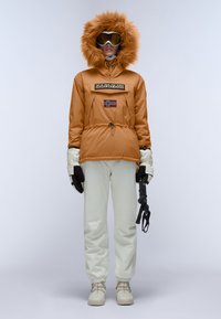 Orange isoleret jakke med pelsforet hætte, snøre i taljen og logo lap; parret med hvide vandtætte bukser og handsker, mens der holds en skistav.