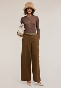 Bruine langemouwen top, brede olijfgroene broek met cargopockets, beige bucket hat, crèmekleurige handtas en beige hoge hakken. Minimalistische accessoires.