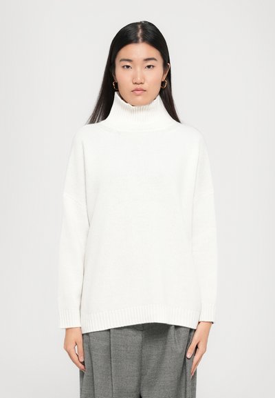 Pull en maille blanche avec col roulé, à la coupe décontractée, avec des poignets et un ourlet côtelés. Modèle portant un pantalon à carreaux gris. Tissu simple et texturé.
