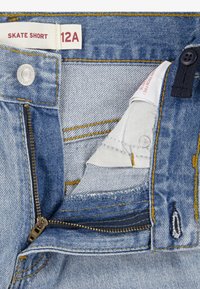 Denim skate shorts med en blå vask, der har gule syninger, sølv lynlås og knaplukning. Indeni er der et mærke med teksten "SKATE SHORT 12A."