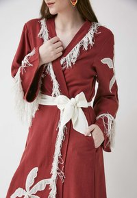 Robe rouge avec des accents de franges blanches et des motifs de dentelle florale. Ceinturée avec un nœud blanc, elle dispose de poches latérales et d'une texture douce.