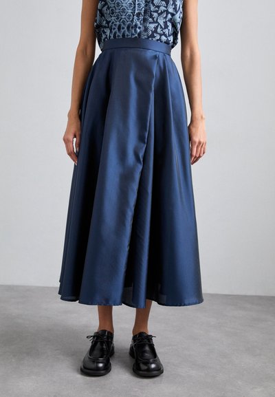Midikleid aus dunkelblauem Satin, mit einem taillierten Bund und plissiertem Design. Kombiniert mit schwarzen Schnürschuhen.