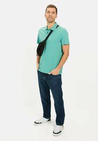 Türkises Poloshirt mit strukturiertem Muster, kombiniert mit dunkelblauen Jeans. Eine schwarze Umhängetasche wird über der Schulter getragen. Weiße Sneaker.