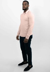 Langärmliges rosa Polohemd mit Kragen, kombiniert mit dunklen passenden Hosen und schwarzen Slippers, getragen von einer stehenden Person.