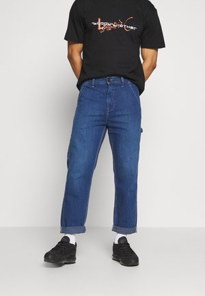 Homme portant un t-shirt noir à motif, un jean bleu retroussé, des chaussettes blanches et des baskets noires, debout devant un fond uni.