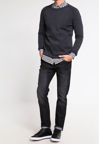 Maglione grigio scuro con maniche raglan sopra una camicia a scacchi bianca e nera, abbinato a jeans scuri e scarpe da ginnastica nere.