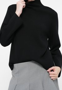 Suéter negro de cuello alto hecho de tela de punto suave, con mangas largas y un corte relajado. Combinado con una falda plisada gris claro.