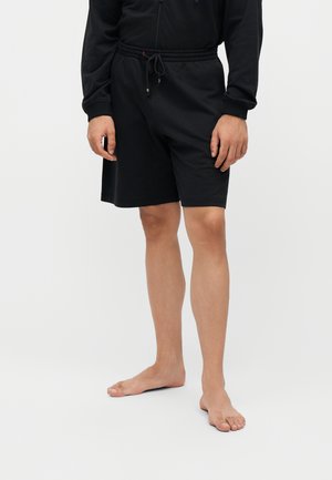 Homme portant un short noir à cordon et un haut noir à manches longues, debout pieds nus sur un fond blanc.