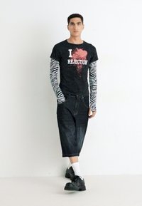 Camiseta negra con el texto "I REJECTION", mangas largas con estampado de cebra, pantalones de mezclilla oscuros y sueltos, y zapatos negros voluminosos con tachuelas.