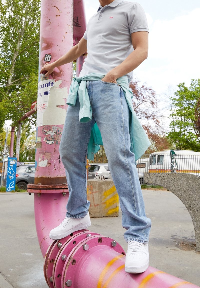 Szary polo z małym logo, jasnoniebieskie dżinsy i białe trampki, stojący na dużej różowej rurze w miejskim otoczeniu na świeżym powietrzu.