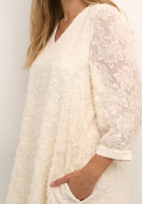 Hvid kjole med struktureret blomsterprint, gennemsigtige ærmer, V-udskæring og sidelommer. Let materiale med bløde udsmykninger.