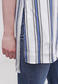 Chemise blanche à rayures verticales bleues et noires, fente latérale, portée avec un jean en denim bleu et un bras détendu le long du corps.