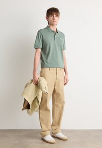 Roheline puuvillane polo särk lühikeste varrukatega ja väikese logoga, koos beezide vabade pükste ja valgete tennisetossudega, hoides kerget jakki.