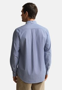 Niebiesko-biała koszula w paski z długim rękawem, kołnierzykiem na guziki, plisą z tyłu i zaokrąglonym dołem. Tkanina bawełniana, luźny krój.