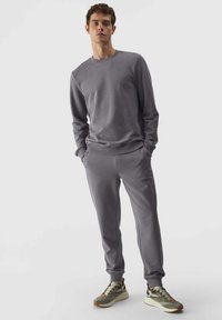Grauer Sweatshirt und passende Jogginghose aus weichem Material, mit gerippten Bündchen und einem dezenten Logo auf der Brust, kombiniert mit Sportschuhen.