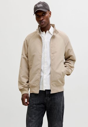 Homme portant une veste beige zippée par-dessus une chemise blanche à rayures avec boutons, un jean noir et une casquette de baseball grise Jack & Jones sur un fond uni.