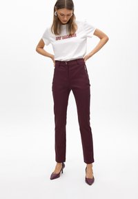 Camiseta blanca con texto rojo, combinada con pantalones burdeos de cintura alta con bolsillos laterales y un diseño ajustado. Modelo lleva tacones morados.