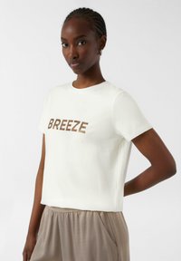 Weißes T-Shirt mit kurzen Ärmeln, auf dem "BREEZE" in strukturierten braunen Buchstaben gedruckt ist, kombiniert mit hellbeigen Hosen. Glatter Stoff, lässige Passform.
