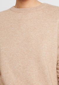 Beige Strickpullover mit rundem Halsausschnitt, der eine weiche Textur und subtile Farbvariationen aufweist. Keine sichtbaren Muster oder Beschläge.