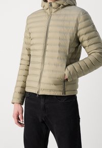 Leichte, olivgrüne, gepolsterte Jacke mit horizontalem Steppmuster, Kapuze, Reißverschlusstaschen und elastischen Bündchen über schwarzen Jeans.