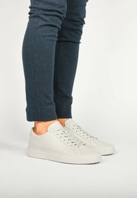 Zapatillas de cuero blancas con una textura suave, punta redonda y un diseño minimalista. Llevadas con pantalones ajustados de color azul oscuro, cortos por el tobillo.