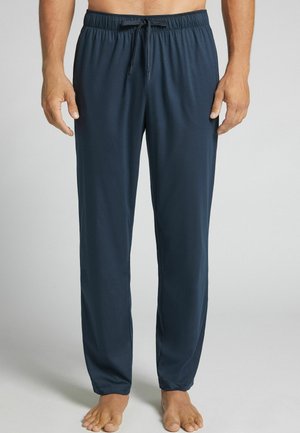 Hombre con pantalones de chándal azul oscuro con cordón, de pie descalzo sobre un fondo claro y sencillo.