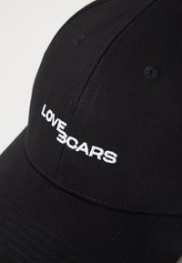 Melna cepure izgatavota no gludas auduma, kuras priekšpusē izšūts balts teksts "LOVE SCARS". Ir izliekta mala un ventilācijas caurumi.