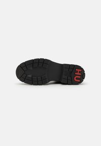 Czarna podeszwa sneakersów z głębokim wzorem bieżnika, teksturowana guma i czerwonym brandingiem "HU". Charakteryzuje się solidnym, płaskim designem dla przyczepności i stabilności.