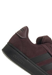 Baskets en suède bordeaux avec semelle en caoutchouc noir, présentant trois rayures noires et un logo en relief sur le talon. Texture lisse avec un design arrondi.