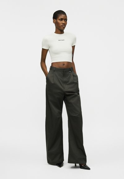 Γυναίκα στέκεται με τα χέρια στις τσέπες, φορώντας λευκό κοντό t-shirt και ψηλόμεσο φαρδύ μαύρο παντελόνι με μαύρες μυτερές γόβες.