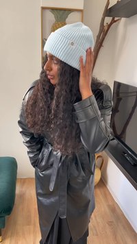 Bonnet en tricot côtelé bleu clair avec un petit logo, porté avec un manteau en cuir noir ceinturé, présentant une coupe décontractée et un style superposé.