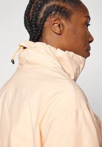 Veste pêche clair avec un col montant, dotée d'un cordon de serrage et de poignets élastiques. Texture lisse, design minimaliste et détails cousus.