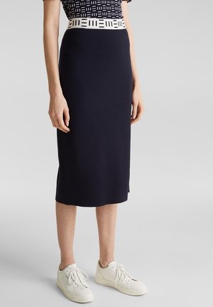 Femme portant une jupe noire au genou avec une ceinture blanche à motif et des baskets blanches, debout devant un fond gris uni.