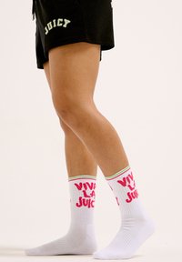 Chaussettes blanches avec le texte "VIVA LA JUICY" en rose et des accents rayés verts et rouges en haut, portées avec un short noir arborant le texte "JUICY" en vert.