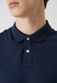 Nahaufnahme einer Person, die ein marineblaues Poloshirt mit Kragen und zwei weißen Knöpfen am Hals trägt.