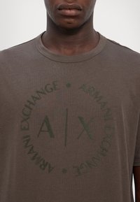Tmavě hnědé tričko z bavlny s kulatým výstřihem, na kterém je zelený kulatý potisk loga obsahující text "Armani Exchange."