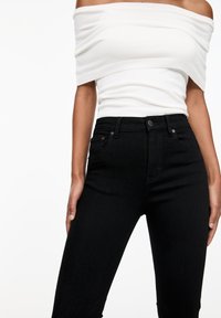 Žena v bílém topu s odhalenými rameny a černých vysokých legínách stojí na pozadí bílé stěny.