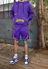Sudadera morada con el logo de "Lakers", bolsillo con cremallera; combinada con pantalones cortos morados y zapatillas blancas con detalles morados. Ambiente al aire libre, suelo de madera.
