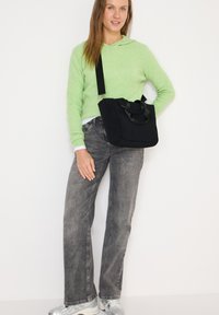 Sac d'épaule en tissu noir avec double poignée et une bandoulière amovible. Associé à un pull vert et un jean gris, sur un fond blanc.