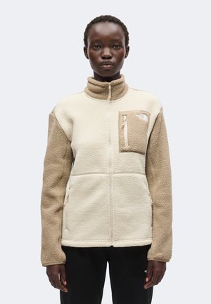 Frau mit kurzen lockigen Haaren trägt eine beige-cremefarbene Fleecejacke mit Reißverschluss und einer Reißverschlusstasche auf der Brust, steht vor einem schlichten Hintergrund.