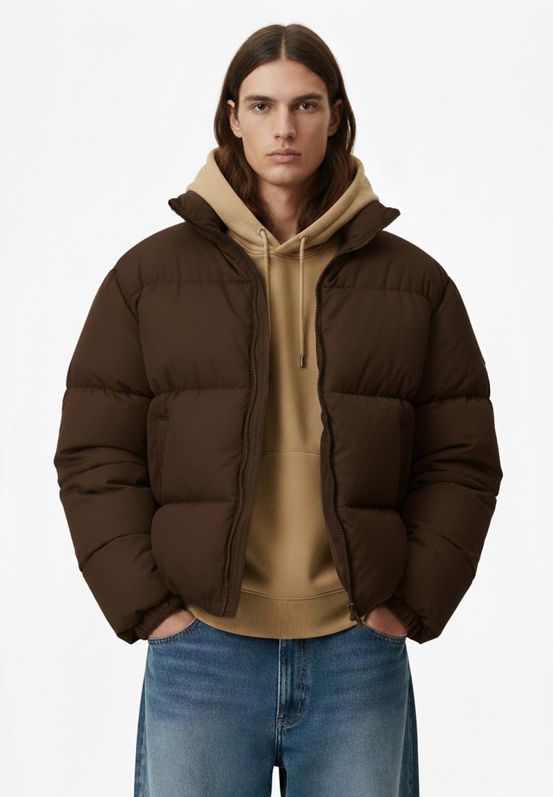 Giacca imbottita marrone con colletto alto, chiusura lampo e due tasche laterali, indossata sopra una felpa beige con cappuccio e jeans azzurri.