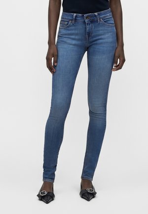 Slim fit blå jeans båret af en person stående med afslappede hænder, parret med sorte spidse flade sko med sølvspænder.