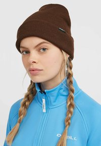 Brun strikket hue med en fold og lille logoetiket; parret med en blå zip-up jakke. Struktureret stof og tætsiddende design.