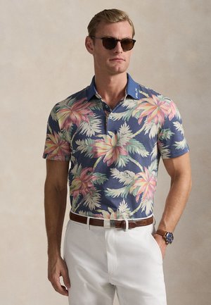 Bărbat purtând ochelari de soare închiși la culoare, o cămașă polo bleumarin cu model floral, pantaloni albi, curea maro și ceas albastru, pe un fundal neutru.